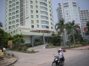 Chữ viết được ghi lại: Trên mặt bên của tòa nhà cao tầng màu vàng nhạt có dòng chữ 'HALONG PEARL Hotel'. Bên trái đường có một biển báo màu vàng nhưng chữ viết trên đó không thể đọc rõ.。Cảnh vật và hoạt động: Bối cảnh là một con đường phía trước một khách sạn lớn ở khu vực đô thị, có thể là thành phố Hạ Long. Tòa nhà chính trong ảnh là khách sạn Halong Pearl. Phía sau là một tòa nhà khách sạn cao tầng khác màu trắng. Khu vực này có nhiều cây cối như cây cọ và cây bụi. Hoạt động chính là hai người đang đi xe máy trên đường. Người ngồi sau đang cầm một bó chổi. Có một chiếc ô tô màu trắng đang đỗ ở bên trái. Lối đi dành cho xe ô tô dẫn lên sảnh chính của khách sạn. Nhiều lá cờ, trong đó có quốc kỳ Việt Nam, đang bay trên các cột cờ.。（日本語訳）。記録された文字：薄黄色の高層ビルの側面には「HALONG PEARL Hotel」という文字があります。道路の左側には黄色い看板がありますが、その上の文字ははっきりと読み取れません。風景と活動：背景は、都市部にある大きなホテルの前の道路で、おそらくハロン市でしょう。写真の主要な建物はハロンパールホテルです。後ろには、別の白い高層ホテルビルがあります。この地域には、ヤシの木や低木など多くの木があります。主な活動は、2人が道路をバイクで走っていることです。後ろに乗っている人はほうきの束を持っています。左側には白い車が1台駐車しています。車用の通路がホテルのメインロビーに通じています。ベトナム国旗を含む多くの旗が旗竿ではためいています。