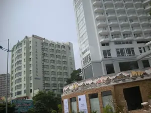 Bức ảnh chụp một góc phố đô thị với các tòa nhà cao tầng, có thể là ở khu vực du lịch Hạ Long. Nổi bật là một tòa nhà màu xanh nhạt với dòng chữ "HALONG PEARL Hotel" được viết dọc theo thân tòa nhà. Bên cạnh đó là một tòa nhà hiện đại cao hơn, màu trắng với rất nhiều ban công. Ở phía trước, một công trình thấp hơn đang được xây dựng hoặc hoàn thiện, có mặt tiền bằng gạch và một bức tranh khảm trên mái mô tả cảnh núi non. Có các biểu ngữ màu trắng treo trên công trình này, một trong số đó ghi "OPENING OCTOBER 1ST", cho thấy sự chuẩn bị cho việc khai trương. Ở góc dưới bên trái, có một biển quảng cáo lớn màu xanh của một công ty có tên "Du Lịch Biển Nhớ". Khung cảnh tổng thể cho thấy sự phát triển của cơ sở hạ tầng du lịch trong khu vực.。（日本語訳）。高層ビルが立ち並ぶ都市の街角を写した写真で、おそらくハロンの観光地でしょう。目立っているのは、建物の側面に沿って「HALONG PEARL Hotel」という文字が縦に書かれた水色の建物です。その隣には、たくさんのバルコニーがある、より高い近代的な白い建物があります。手前には、建設中または完成間近の低い建物があり、レンガ造りのファサードと、山々の風景を描いた屋根のモザイク画が特徴です。この建物には白い横断幕が掲げられており、そのうちの一つには「OPENING OCTOBER 1ST」と書かれていて、開業準備が進められていることがわかります。左下隅には、「Du Lịch Biển Nhớ」という名前の会社の大きな青い看板があります。全体的な光景は、この地域の観光インフラの発展を示しています。