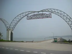 Bức ảnh chụp một cổng chào lớn bằng kim loại có kết cấu dạng vòm, đây là lối vào Khu du lịch Đảo Tuần Châu ở khu vực Hạ Long. Trên cổng chào có treo một tấm biển lớn, nhưng dòng chữ trên đó bị ngược trong ảnh. Nếu đọc xuôi, nội dung trên biển là: "TỈNH QUẢNG NINH", "KHU DU LỊCH ĐẢO TUẦN CHÂU", và "KÍNH CHÀO QUÝ KHÁCH". Con đường nhựa bên dưới cổng chào dẫn thẳng ra một con đường dài vượt biển để vào đảo. Hai bên con đường là biển cả, và ở phía xa có thể thấy những hòn đảo hoặc dãy núi mờ ảo trong sương. Bên phải đường có một bốt gác nhỏ màu xanh và một vài người cùng xe máy đang đỗ. Dọc theo con đường vượt biển có lắp đặt các cột đèn đường. Khung cảnh cho thấy một công trình hạ tầng du lịch ven biển.。（日本語訳）。写真には、アーチ構造の大きな金属製のウェルカムゲートが写っており、これはハロン地区にあるトゥアンチャウ島観光エリアへの入り口です。ウェルカムゲートには大きな看板が掲げられていますが、その上の文字は写真では反転しています。順方向に読むと、看板の内容は「クアンニン省」、「トゥアンチャウ島観光エリア」、そして「お客様を歓迎します」です。ゲートの下のアスファルトの道は、島へ入るために海を渡る長い道へとまっすぐ続いています。道の両側は海で、遠くには霧の中にぼんやりと島々や山脈が見えます。道路の右側には小さな緑色の警備詰所があり、数人とバイクが停まっています。海を渡る道に沿って街灯が設置されています。この光景は、沿岸の観光インフラ施設を示しています。