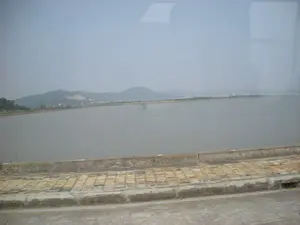 Bức ảnh chụp một khung cảnh ven biển, có thể được chụp từ một phương tiện đang di chuyển như ô tô hoặc xe buýt. Ở phía trước là một phần của con đường hoặc cây cầu bằng bê tông. Sát lề đường là một khu vực được chia thành nhiều ô vuông, trông giống như sân phơi (có thể là phơi hải sản) hoặc ruộng muối. Phần chính của bức ảnh là một vùng nước rộng lớn, mặt nước phẳng lặng, có khả năng là một đầm nuôi trồng thủy sản hoặc một phần của vịnh. Giữa vùng nước có một công trình nhỏ giống như một chòi canh được dựng trên các cọc gỗ. Ở phía xa là những dãy đồi núi bao quanh. Bầu trời có màu xám và mờ ảo do sương mù. Không có văn bản hay biển hiệu nào có thể nhận dạng được trong ảnh.。（日本語訳）。写真は沿岸の風景を写しており、自動車やバスのような移動中の乗り物から撮影された可能性があります。手前には、コンクリート製の道路または橋の一部があります。道端に沿って、多くの四角い区画に分けられたエリアがあり、干し場（海産物を干している可能性あり）または塩田のように見えます。写真の主要部分は広大な水域で、水面は穏やかであり、水産養殖の池または湾の一部である可能性があります。水域の中央には、木製の杭の上に建てられた見張り小屋のような小さな建造物があります。遠くには、周囲を囲む丘や山々の連なりがあります。空は灰色で、霧のために霞んでいます。写真の中には、識別できる文字や看板はありません。