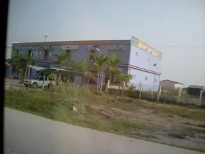 Trên mặt tiền của một tòa nhà hai tầng sơn màu tím nhạt có một biển hiệu lớn với dòng chữ: "TRANG TRÍ NỘI THẤT & VẬT LIỆU XÂY DỰNG THIÊN ÂN". Ngoài ra còn có một tấm biển quảng cáo khác ở bên hông tòa nhà nhưng chữ bị mờ, không đọc được.。Bối cảnh là một khu vực ven đường, có thể là ở ngoại ô. Tòa nhà này là một cửa hàng kinh doanh vật liệu xây dựng và đồ trang trí nội thất. Phía trước tòa nhà có một mái che đơn giản, dưới đó có một chiếc xe ô tô bán tải màu bạc đang đỗ. Xung quanh tòa nhà được trồng một vài cây cọ. Ở phía trước, giữa tòa nhà và con đường nhựa là một khu đất trống có cỏ mọc. Không có hoạt động nào của con người được ghi lại một cách rõ ràng.。（日本語訳）。薄紫色に塗られた2階建ての建物の正面には、「ティエンアン内装＆建材」という文字が書かれた大きな看板があります。また、建物の側面にも別の広告看板がありますが、文字がかすれていて読めません。背景は道端のエリアで、おそらく郊外でしょう。この建物は建材と内装品を販売する店です。建物の前には簡単な屋根があり、その下には銀色のピックアップトラックが1台駐車しています。建物の周りには数本のヤシの木が植えられています。前方、建物とアスファルトの道路の間には、草が生えている空き地があります。人間の活動は明確には記録されていません。