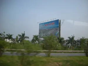 Bức ảnh chụp cảnh quan ven đường từ một phương tiện đang di chuyển. Ở trung tâm bức ảnh là một tấm biển quảng cáo lớn. Trên biển quảng cáo có dòng chữ: "ĐÔ THỊ MỚI NAM TỪ SƠN" và "Hội tụ những giá trị Châu Âu trong lòng Hà Nội - Bắc Ninh". Tấm biển này quảng cáo cho một dự án khu đô thị mới, với hình ảnh phối cảnh các tòa nhà và nhà ở hiện đại. Phía trước và xung quanh tấm biển là cây cối xanh tươi, bao gồm cả cây cọ, và một con đường có hàng rào chắn. Phía sau tấm biển có thể nhìn thấy một vài ngôi nhà và biệt thự. Bầu trời có màu xám và u ám. Hình ảnh ở phía trước bị mờ do chuyển động.。（日本語訳）。移動中の乗り物から撮影された道端の風景写真です。写真の中央には、大きな広告看板があります。広告看板には、「ĐÔ THỊ MỚI NAM TỪ SƠN」および「Hội tụ những giá trị Châu Âu trong lòng Hà Nội - Bắc Ninh」（ハノイ-バクニンの中心にヨーロッパの価値が集結）という文言があります。この看板は、近代的なビルや住宅の完成予想図とともに、新都市区プロジェクトを宣伝しています。看板の前と周りには、ヤシの木を含む青々とした木々や、ガードレールのある道路があります。看板の後ろには、いくつかの家や別荘が見えます。空は灰色で曇っています。前景の画像は動きによってぼやけています。