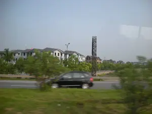 Bức ảnh được chụp từ một phương tiện đang di chuyển, cho thấy khung cảnh dọc một con đường lớn hoặc đường cao tốc. Do hiệu ứng mờ của chuyển động ở tiền cảnh, có thể thấy rõ đây là góc nhìn từ bên trong xe. Một chiếc ô tô màu đen đang di chuyển trên làn đường song song. Ở hậu cảnh là một dãy nhà liền kề nhiều tầng, có vẻ là một khu đô thị mới. Các ngôi nhà có tường trắng và mái ngói sẫm màu. Trên nóc một ngôi nhà có cắm lá cờ Việt Nam. Xen kẽ là cây xanh và một vài công trình khác, bao gồm một kết cấu thép dạng lưới cao và một số ngôi nhà đang trong quá trình xây dựng. Không có văn bản hay chữ viết nào có thể nhận dạng được trong hình ảnh.。（日本語訳）。前景のモーションブラー効果により、これが車内からの視点であることがはっきりとわかります。並行する車線を黒い車が走行しています。背景には、新興住宅地と思われる多階建ての連続住宅が並んでいます。家々は白い壁と暗い色の瓦屋根です。一軒の家の屋根にはベトナムの国旗が掲げられています。緑の木々や、高い格子状の鉄骨構造物、建設中の家々など、いくつかの他の建造物が点在しています。画像内には識別可能なテキストや文字はありません。