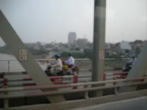 Bức ảnh chụp cảnh giao thông trên một cây cầu giàn thép. Nhiều người đang điều khiển xe máy đi qua cầu. Một người lái xe đội mũ bảo hiểm màu xanh. Phía sau họ, một người khác mặc áo sơ mi trắng cũng đang lái xe máy. Cây cầu có kết cấu bằng kim loại, bắc qua một con sông. Ở phía xa là quang cảnh của một thành phố với nhiều tòa nhà. Trên một trụ cầu bằng thép có một biển báo gồm hai ô vuông nhỏ viền đỏ, xếp chồng lên nhau. Chữ và số bên trong rất mờ, khó đọc, có vẻ như là 'W17' và 'W24'.。（日本語訳）。鋼鉄のトラス橋の上の交通風景を撮影した写真です。多くの人がバイクを運転して橋を渡っています。ある運転手は青いヘルメットをかぶっています。彼らの後ろでは、別の人が白いシャツを着てバイクを運転しています。橋は金属構造で、川に架かっています。遠くには、多くの建物がある街の景色が見えます。鋼鉄の橋脚の一つに、赤枠の小さな四角が二つ上下に重なった標識があります。中の文字と数字は非常にぼやけていて読みにくく、「W17」と「W24」のように見えます。