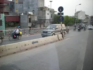 Cảnh quan giao thông trên một cây cầu hoặc cầu vượt tại một khu vực đô thị. Nhiều người đang điều khiển xe máy và ô tô trên đường. Một số người đi xe máy đội mũ bảo hiểm và đeo khẩu trang. Có một chiếc xe máy chở hai người, người ngồi sau xách một chiếc túi màu vàng. Làn đường được phân chia bởi một dải phân cách bằng bê tông, trên đó có vẽ một mũi tên màu đen chỉ hướng sang trái. Bên đường có một cột biển báo giao thông với hai biển báo hình tròn màu xanh lam nhưng không rõ nội dung. Phía sau là những ngôi nhà nhiều tầng san sát nhau, một số có bồn nước inox trên mái nhà. Lan can cầu có màu trắng và đỏ.。（日本語訳）。都市部の橋または高架橋の交通風景。多くの人々が道路でバイクや自動車を運転している。バイクに乗っている人の中には、ヘルメットをかぶり、マスクをしている人もいる。2人乗りのバイクが1台あり、後ろに乗っている人は黄色いバッグを持っている。車線はコンクリートの中央分離帯で分けられており、その上には左を指す黒い矢印が描かれている。道路の脇には交通標識のポールがあり、青い円形の標識が2つ付いているが、内容は不明瞭である。後方には多層階の家々が密集しており、中には屋上にステンレス製の貯水タンクがある家もある。橋の欄干は白と赤である。