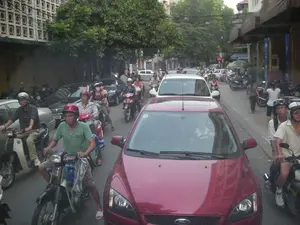 Bức ảnh chụp cảnh giao thông đông đúc trên một con đường trong thành phố tại Việt Nam. Dòng xe cộ gồm rất nhiều xe máy và ô tô đang chen chúc nhau, có vẻ như đang trong tình trạng ùn tắc hoặc di chuyển rất chậm. Ở cận cảnh là một chiếc ô tô màu đỏ hiệu Ford. Rất nhiều người đang điều khiển xe máy, và hầu hết họ đều đội mũ bảo hiểm. Hai bên đường là các tòa nhà nhiều tầng và cây xanh. Tòa nhà bên trái có mặt tiền với các khối bê tông hoa văn. Các tòa nhà bên phải có mái hiên, trông giống như mặt tiền của các cửa hàng hoặc khách sạn. Một vài người đang đi bộ trên vỉa hè bên phải. Ở phía xa có một biển báo giao thông hình tròn màu đỏ với vạch trắng ngang, là biển báo cấm đi ngược chiều.。（日本語訳）。ベトナムのある都市の道路での混雑した交通風景を撮影した写真です。車の流れは、多くのバイクと自動車で構成されており、互いにひしめき合っています。渋滞しているか、非常にゆっくりと移動しているようです。前景にはフォード製の赤い車があります。多くの人がバイクを運転しており、そのほとんどがヘルメットを着用しています。道路の両側には、複数階建ての建物と緑の木々があります。左側の建物は、模様の入ったコンクリートブロックでできたファサードをしています。右側の建物には日よけがあり、店舗やホテルの正面のように見えます。右側の歩道では数人が歩いています。遠くには、白い横線が入った赤色の円形交通標識があります。これは進入禁止の標識です。