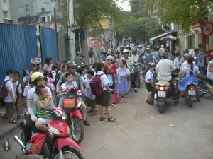 Đây là một khung cảnh đông đúc trên một con ngõ hoặc đường phố nhỏ ở Việt Nam, có vẻ như là giờ tan trường. Có một tấm biển nhỏ màu đỏ với chữ trắng ghi "LỚP 3M". Ở phía bên phải, có một biển báo giao thông hình tròn, viền đỏ, nền trắng với hình ô tô màu đen bị gạch chéo, đây là biển báo cấm ô tô. Địa điểm có thể là khu vực trước cổng một trường tiểu học, thể hiện qua sự hiện diện của rất nhiều học sinh mặc đồng phục (áo trắng, quần/váy tối màu) và đeo cặp sách. Hoạt động chính là các bậc phụ huynh đang dùng xe máy để đến đón con em mình. Cảnh tượng khá náo nhiệt và có phần hỗn loạn với nhiều người và xe máy chen chúc nhau trên con đường. Một số người lớn và trẻ em ngồi trên xe máy có đội mũ bảo hiểm. Bên trái bức ảnh là một hàng rào bằng tôn màu xanh. Phía sau là các tòa nhà dân cư và cây xanh.。（日本語訳）。これはベトナムの路地または小さな通りでの混雑した光景で、下校時間だと思われます。「LỚP 3M」と白文字で書かれた小さな赤い看板があります。右側には、赤い縁取り、白い背景に黒い自動車の絵が斜線で消された円形の交通標識があります。これは自動車通行止めの標識です。場所は小学校の校門前エリアかもしれません。制服（白いシャツ、暗い色のズボン/スカート）を着てランドセルを背負った多くの生徒がいることからそれが分かります。主な活動は、保護者たちがバイクで自分の子供を迎えに来ていることです。多くの人々やバイクが道にひしめき合っており、光景はかなり賑やかで、やや混沌としています。バイクに乗っている大人や子供の中には、ヘルメットを着用している人もいます。写真の左側には、青いトタンのフェンスがあります。背景には住宅ビルと緑の木々があります。