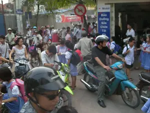 Bức ảnh ghi lại cảnh tượng giao thông đông đúc trên một con đường ở Việt Nam, có thể là vào giờ tan học. Rất nhiều xe máy đang dừng hoặc di chuyển chậm, tạo nên cảnh ùn tắc. Trong ảnh có nhiều người lớn và trẻ em. Các em học sinh mặc đồng phục gồm áo sơ mi trắng, quần hoặc váy màu xanh, một số em đeo khăn quàng đỏ và mang ba lô. Nhiều người lớn đang điều khiển xe máy và đội mũ bảo hiểm, có vẻ như là phụ huynh đang đón con. Bên phải đường có một cửa hàng sửa chữa xe máy với biển hiệu dọc màu xanh. Trên biển hiệu có logo của hãng "Mobil" và các dòng chữ "Bảo Dưỡng", "Sửa Chữa", "Tất Cả". Phía trên, có một biển báo giao thông hình tròn, nền trắng, viền đỏ với biểu tượng ô tô bị gạch chéo, đây là biển báo cấm ô tô. Ở phía sau, có thể thấy một băng rôn màu đỏ và một biển báo đường màu xanh nhưng không thể đọc rõ nội dung.。（日本語訳）。この写真はベトナムのある道路での混雑した交通風景を記録したもので、おそらく下校時間帯でしょう。非常に多くのバイクが停止またはゆっくりと移動しており、渋滞の光景を作り出しています。生徒たちは白いシャツ、青いズボンまたはスカートからなる制服を着ており、一部の生徒は赤いスカーフを身に着け、バックパックを背負っています。多くの大人がバイクを運転しヘルメットを着用しており、子供を迎えに来た保護者のようです。道路の右側には、青い縦長の看板があるバイク修理店があります。看板には「モービル」社のロゴと、「Bảo Dưỡng」、「Sửa Chữa」、「Tất Cả」という文字があります。その上には、白い背景に赤い縁取りの円形をした交通標識があり、斜線が引かれた自動車のシンボルが描かれています。これは自動車進入禁止の標識です。後方には、赤い横断幕と青い道路標識が見えますが、内容ははっきりと読み取れません。