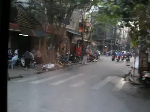 Hình ảnh ghi lại một cảnh đường phố ở Việt Nam. Con đường có nhiều người đang di chuyển bằng xe máy. Trên mặt đường có vạch kẻ sang đường cho người đi bộ. Dọc hai bên đường là các tòa nhà, cây xanh và hệ thống dây điện chằng chịt phía trên. Ở vỉa hè bên trái, một nhóm người đang tụ tập, một số ngồi trên ghế nhựa nhỏ. Có một chiếc xe đạp màu vàng và một vài chiếc xe máy đang đậu gần đó. Có các biển hiệu trên các tòa nhà nhưng chữ bị mờ và không thể đọc được do chất lượng hình ảnh.。（日本語訳）。画像はベトナムのある街の風景を捉えています。道路では多くの人々がバイクで移動しています。路面には歩行者用の横断歩道の線が引かれています。道路の両脇には建物、緑の木々、そして上部には複雑に絡み合った電線があります。左側の歩道では、一群の人々が集まっており、何人かは小さなプラスチックの椅子に座っています。近くには黄色い自転車1台と数台のバイクが停まっています。建物には看板がありますが、画質が悪いため文字がぼやけていて読むことができません。