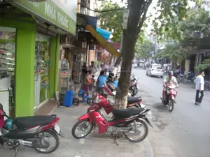 Bức ảnh chụp một cảnh đường phố ban ngày ở Việt Nam.。**Chữ viết:** Trên một cửa hàng có mặt tiền màu xanh lá cây, có một biển hiệu lớn với dòng chữ "NHÀ THUỐC" và chữ "PHARMACY" nhỏ hơn ở trên.。**Bối cảnh và hoạt động:** Địa điểm là một con phố với các cửa hàng san sát nhau. Trọng tâm của bức ảnh là một nhà thuốc có cửa kính, bên trong có thể thấy các kệ chứa đầy thuốc. Trên vỉa hè phía trước, một nhóm người đang ngồi trên những chiếc ghế nhựa nhỏ màu xanh và đỏ, có vẻ như đang nghỉ ngơi hoặc trò chuyện. Nhiều xe máy được dựng trên vỉa hè, nổi bật là hai chiếc ở gần máy ảnh. Dưới lòng đường, một người đàn ông đội mũ bảo hiểm đang lái xe máy màu đỏ chở theo một đứa trẻ ngồi phía trước. Phía sau họ là một chiếc ô tô màu trắng và nhiều phương tiện khác đang lưu thông. Một người đàn ông mặc áo trắng đang đi bộ bên lề đường. Cây cối được trồng dọc theo vỉa hè, tạo nên khung cảnh đặc trưng của đường phố đô thị.。（日本語訳）。この写真はベトナムの昼間の街の風景を撮影したものです。**文字:** 緑色のファサードを持つ店舗の上に、「NHÀ THUỐC」という大きな文字と、その上に小さく「PHARMACY」と書かれた看板があります。**背景と活動:** 場所は、店舗が隣接して立ち並ぶ通りです。写真の中心はガラス戸の薬局で、中には薬がぎっしり詰まった棚が見えます。手前の歩道では、数人のグループが青と赤の小さなプラスチック製の椅子に座っており、休憩しているか、おしゃべりをしているようです。歩道には多くのバイクが停められており、特にカメラに近い2台が目立ちます。車道では、ヘルメットをかぶった男性が赤いバイクを運転しており、前には子供が一人乗っています。彼らの後ろには白い車と、その他多くの車両が通行しています。白いシャツを着た男性が道端を歩いています。歩道に沿って木々が植えられており、都市の街路に特徴的な風景を作り出しています。