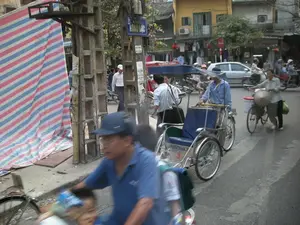 Bức ảnh chụp một cảnh đường phố nhộn nhịp ở Việt Nam. Có một biển báo đường phố màu xanh ghi chữ "PHỐ HÀNG BẠC". Trên cột điện còn có một biển báo giao thông hình tròn nền trắng viền đỏ (biển báo cấm đi ngược chiều). Trên thân một chiếc xe xích lô có thể đọc được chữ "VINASUN" và số "3". Các biển hiệu và giấy dán khác trên cột điện và các tòa nhà không thể đọc rõ nội dung.。Địa điểm là một con phố đô thị với những tòa nhà nhiều tầng san sát nhau và hệ thống dây điện chằng chịt. Hoạt động chính là giao thông tấp nập với nhiều loại phương tiện. Một người đàn ông đang đạp một chiếc xích lô (cyclo) không có khách, có lẽ đang tìm khách. Một người phụ nữ đội nón lá sau lưng đang dắt một chiếc xe đạp có chở một cái xô màu đỏ và một chiếc túi, có thể là người bán hàng rong hoặc đi chợ về. Ở phía trước, một người đàn ông mặc áo phông xanh và đội mũ lưỡi trai đang lái xe, hình ảnh bị nhòe do chuyển động. Nhiều người khác đang đi xe máy và đi bộ trên đường. Các vật thể đáng chú ý bao gồm xe xích lô, xe máy, ô tô, xe đạp, nón lá, và một tấm bạt lớn có sọc màu xanh, đỏ, trắng che phủ một khu vực bên lề đường.。（日本語訳）。この写真はベトナムの賑やかな街の風景を撮影したものです。「PHỐ HÀNG BẠC」と書かれた青い道路標識があります。電柱には、白地に赤枠の円形交通標識（一方通行出口の進入禁止標識）もあります。シクロの車体には「VINASUN」という文字と「3」という数字が読み取れます。電柱や建物にある他の看板や貼り紙は、内容がはっきりと読み取れません。場所は、多層階の建物が密集し、電線が複雑に張り巡らされた都市の通りです。主な活動は、さまざまな種類の乗り物による混雑した交通です。一人の男性が客の乗っていないシクロを漕いでおり、おそらく客を探しているところでしょう。背中にノンラーをかぶった女性が、赤いバケツと鞄を乗せた自転車を押しており、露天商か市場からの帰りかもしれません。手前では、青いTシャツを着て野球帽をかぶった男性が運転していますが、動きによって画像がぼやけています。他にも多くの人々がバイクに乗ったり、道を歩いたりしています。注目すべき物には、シクロ、バイク、自動車、自転車、ノンラー、そして道端の一角を覆う青、赤、白の縞模様の大きな防水シートが含まれます。