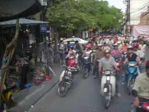 Bức ảnh chụp một con phố đông đúc và nhộn nhịp tại một thành phố ở Việt Nam. Con đường chật cứng người và phương tiện, chủ yếu là xe máy, tạo nên cảnh tượng có thể là tắc đường. Hầu hết những người điều khiển xe máy đều đội mũ bảo hiểm. Ở phía sau có thể thấy một chiếc ô tô màu bạc. Bên trái đường, trên vỉa hè, có một quầy hàng tạm bợ với mái hiên kẻ sọc xanh trắng, được chống đỡ bằng các cọc tre. Một người phụ nữ đang cúi xuống sắp xếp hàng hóa được bày bán, bao gồm nhiều mặt hàng treo lủng lẳng. Phía trước quầy hàng có vài chiếc ghế nhựa nhỏ. Dọc con phố là những tòa nhà cao tầng và cây xanh. Có một biển báo đường phố màu xanh lam với chữ trắng nhưng không thể đọc rõ nội dung. Phía xa hơn có một biển báo giao thông hình tròn. Hệ thống dây điện chằng chịt vắt ngang phía trên, một hình ảnh quen thuộc ở các đô thị Việt Nam.。（日本語訳）。この写真はベトナムのある都市の、混雑して賑やかな通りを撮影したものです。道路は人と乗り物、主にバイクで埋め尽くされており、交通渋滞かもしれない光景を作り出しています。バイクを運転している人のほとんどはヘルメットを着用しています。後方には銀色の自動車が見えます。道路の左側、歩道の上には、青と白の縞模様の軒先テントがあり、竹の棒で支えられた仮設の屋台があります。一人の女性が、ぶら下がっている多くの商品を含む、販売用の商品を並べるためにかがんでいます。屋台の前にはいくつかの小さなプラスチックの椅子があります。通り沿いには高層ビルや緑の木々が並んでいます。青地に白文字の道路標識がありますが、内容ははっきりと読み取れません。さらに遠くには円形の交通標識があります。頭上には電線が複雑に張り巡らされており、ベトナムの都市部ではおなじみの光景です。