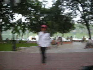 Bức ảnh bị mờ do chuyển động, ghi lại khung cảnh tại một công viên hoặc một con đường đi dạo ven bờ hồ hoặc vịnh. Ở tiền cảnh, một người đội mũ bảo hiểm màu đỏ, mặc áo khoác trắng và quần tối màu đang di chuyển. Vỉa hè được lát bằng gạch đỏ. Ở hậu cảnh, có nhiều cây xanh lớn và một vùng nước. Có thể thấy một vài người khác đang ngồi nghỉ ngơi dọc theo bờ kè hoặc trên ghế đá. Do ảnh bị nhòe nên không thể đọc được bất kỳ văn bản hay biển hiệu nào.。（日本語訳）。写真は手ブレでぼやけており、公園、あるいは湖か湾のほとりにある遊歩道の風景を捉えています。前景には、赤いヘルメットをかぶり、白いジャケットと暗い色のズボンを履いた人物が移動しています。歩道は赤いレンガで舗装されています。背景には、多くの大きな緑の木々と水辺があります。他に数人が護岸沿いや石のベンチに座って休んでいるのが見えます。