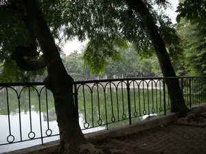 Đây là một khung cảnh ven hồ, có thể là trong một công viên ở Hà Nội. Ở phía trước là một lối đi bộ lát gạch. Một hàng rào sắt màu đen, được trang trí bằng các họa tiết hình vòng cung và hình tròn, chạy dọc theo bờ hồ. Những cây cổ thụ lớn với tán lá xanh um tùm mọc ngay cạnh hàng rào, đổ bóng mát xuống lối đi. Mặt hồ phẳng lặng và có màu xanh lục. Nhìn qua hàng rào, ở phía bên kia hồ, có thể thấy nhiều cây xanh và một vài công trình xây dựng ở phía xa. Không có hoạt động cụ thể nào của con người được ghi lại, khung cảnh rất yên tĩnh. Không có chữ viết nào có thể đọc được trong ảnh.。（日本語訳）。これは湖畔の風景で、おそらくハノイの公園の中でしょう。手前にはレンガ敷きの遊歩道があります。弧と円の模様で装飾された黒い鉄の柵が、湖岸に沿って続いています。青々と茂る葉を持つ大きな古木が柵のすぐそばに生えており、遊歩道に涼しい影を落としています。湖面は穏やかで、緑色をしています。柵越しに湖の向こう側を見ると、たくさんの緑の木々と、遠くにいくつかの建物が見えます。特定の人の活動は記録されておらず、風景はとても静かです。写真の中に読み取れる文字はありません。