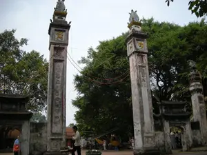 Bức ảnh chụp cổng vào của một khu di tích lịch sử hoặc tôn giáo, có thể là một ngôi đình hoặc chùa ở khu vực Hà Nội. Nổi bật nhất là hai cột đồng trụ (trụ biểu) lớn bằng đá, trông cũ kỹ và cổ kính. Trên thân hai cột trụ có khắc các câu đối bằng Chữ Hán hoặc Chữ Nôm theo chiều dọc, nhưng các ký tự đã mờ và khó đọc. Ngoài ra còn có các bức phù điêu nhỏ hình linh vật được sơn vàng. Trên đỉnh mỗi cột là các họa tiết trang trí điêu khắc. Giữa hai cột trụ có giăng một ngôi sao lớn màu vàng, đây có thể là vật trang trí cho một dịp lễ hội như Tết Trung Thu. Phía sau các cột trụ là một cổng phụ có mái và tường bao, cũng mang nét kiến trúc cổ. Xung quanh là không gian rộng với nhiều cây xanh. Có một vài người đang đi lại trong khu vực. Ở phía sau, có thể thấy một chiếc xe công trình màu vàng, cho thấy có thể đang có hoạt động tu sửa hoặc xây dựng gần đó.。（日本語訳）。ハノイ地域にある歴史遺跡または宗教施設の入り口の門を撮った写真で、おそらくは亭（てい）か寺でしょう。最も目立つのは、石でできた2本の大きな同柱（標柱）で、古びて風格があるように見えます。2本の柱の胴体には、漢字またはチュノムによる対句が縦に刻まれていますが、文字は薄れて読みにくくなっています。その他に、金色に塗られた霊獣の形の小さな浮き彫りもあります。各柱の頂上には、彫刻された装飾模様があります。2本の柱の間には大きな黄色い星が張られており、これは中秋節のような祭りの機会のための装飾物かもしれません。柱の後ろには、屋根と囲い壁のある副門があり、これもまた古い建築様式を帯びています。周囲は多くの緑の木々がある広い空間です。その地域内を何人かの人が歩き回っています。後方には、黄色い工事車両が一台見え、その近くで修復または建設活動が行われている可能性があることを示しています。
