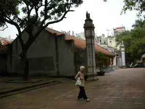 Bối cảnh là sân của một ngôi chùa hoặc đình cổ ở Hà Nội. Một bà cụ tóc bạc, mặc áo màu nâu nhạt và quần đen, đang đi bộ qua khoảng sân rộng lát gạch đỏ. Bà đang xách một chiếc túi ni lông màu trắng. Bên trái có một cây cổ thụ lớn. Kiến trúc của ngôi chùa mang nét cổ kính với tường xám và mái lợp ngói vảy cá màu đỏ cam. Trong sân có các trụ biểu bằng đá, trên đó có khắc các câu đối bằng chữ Hán. Cột trụ ở gần có thể đọc được dòng chữ: "地涌金蓮中寶臺" (Địa dũng kim liên trung bảo đài). Phía sau là các công trình khác của ngôi chùa và thấp thoáng những ngôi nhà dân sinh hiện đại. Một chiếc xe máy đang đỗ ở phía xa bên phải.。（日本語訳）。背景はハノイのある古いお寺か亭（てい）の庭である。白髪のおばあさんが、薄茶色の上着と黒いズボンを履き、赤レンガが敷かれた広い庭を歩いている。彼女は白いビニール袋を提げている。左側には大きな古木がある。お寺の建築は、灰色の壁と赤みがかったオレンジ色の魚の鱗のような瓦で葺かれた屋根で、古風な趣を呈している。庭には石の柱があり、その上には漢文の対句が刻まれている。近くの柱には「地涌金蓮中寶臺」（Địa dũng kim liên trung bảo đài）という文字が読み取れる。遠くの右側にはバイクが一台停まっている。