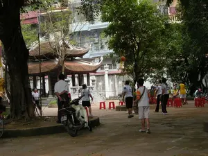 Bối cảnh là một không gian công cộng ngoài trời, có thể là sân của một ngôi đình hoặc chùa ở Hà Nội, với nhiều cây cổ thụ lớn. Ở hậu cảnh, có một công trình kiến trúc truyền thống với mái ngói cong đặc trưng, nằm xen kẽ giữa các tòa nhà dân sinh hiện đại. Một nhóm người đang tụ tập trên sân. Nhiều người trong số họ mặc trang phục thể thao và đang chơi một trò chơi, có thể là bóng rổ, được thể hiện qua hình ảnh một quả bóng rổ đang bay trên không. Xung quanh có nhiều ghế nhựa màu đỏ để ngồi. Ở bên trái, một người đàn ông mặc áo sơ mi trắng đang đứng cạnh một chiếc xe máy tay ga đang đỗ. Không có văn bản nào có thể đọc được từ các biển báo hay công trình kiến trúc trong ảnh.。（日本語訳）。背景は屋外の公共スペースで、ハノイにある亭か寺院の庭かもしれず、大きな古木が多数あります。背景には、特徴的な反りのある瓦屋根を持つ伝統的な建築物があり、現代的な民間の建物の間に点在しています。一団の人々が庭に集まっています。彼らの多くはスポーツウェアを着てゲームをしています。それはバスケットボールの可能性があり、空中に飛んでいるバスケットボールのイメージによって示されています。周りには座るための赤いプラスチックの椅子がたくさんあります。左側では、白いシャツを着た男性が、駐車しているスクーターの隣に立っています。写真の中の看板や建築物からは、読み取れる文字はありません。