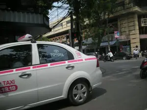 Bức ảnh chụp một cảnh đường phố nhộn nhịp tại một thành phố, có thể là Hà Nội. Đối tượng chính trong ảnh là một chiếc xe taxi màu trắng có sọc trang trí màu hồng đang di chuyển trên đường. Trên thân xe có các chữ và số có thể đọc được: "TAXI", một phần số điện thoại "...19.19", và số "150" trong một vòng tròn màu hồng. Cửa kính sau của xe có dán logo "TOYOTA". Bối cảnh xung quanh là một con đường đô thị với các tòa nhà cao tầng, cây xanh và một hệ thống dây điện, dây cáp chằng chịt trên không. Hoạt động giao thông đang diễn ra với nhiều xe máy và ô tô khác trên đường. Có thể thấy một người đang lái xe máy đội mũ bảo hiểm và một vài người đang đứng trên vỉa hè ở phía xa. Một cột đèn giao thông đang hiển thị màu đỏ.。（日本語訳）。写真は、ある都市、おそらくハノイの賑やかな街の風景を撮影したものです。写真の主な被写体は、道路を走行している、ピンク色の装飾的なストライプが入った白いタクシーです。車体には、「TAXI」、電話番号の一部「...19.19」、そしてピンク色の円の中に「150」という数字が読み取れます。車の後部窓には「TOYOTA」のロゴが貼られています。周囲の背景は、高層ビル、緑の木々、そして上空に張り巡らされた電線やケーブルのシステムがある都市の道路です。道路上では多くのバイクや他の車が行き交い、交通活動が行われています。ヘルメットをかぶってバイクを運転している人や、遠くの歩道に立っている数人の人々が見えます。信号機が赤色を表示しています。