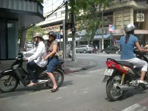 Bối cảnh là một con đường nhựa trong thành phố vào ban ngày, có nhiều người và phương tiện đang tham gia giao thông. Ở phía trước, có hai chiếc xe máy. Chiếc xe máy màu đen bên trái do một người đàn ông mặc áo sơ mi trắng, quần tối màu điều khiển, phía sau chở một người phụ nữ mặc áo phông nâu và váy xanh. Cả hai đều đội mũ bảo hiểm. Người phụ nữ có đeo một chiếc túi xách hoa văn. Chiếc xe máy màu đỏ bên phải do một người phụ nữ mặc áo phông xanh, quần trắng điều khiển một mình. Cô ấy đội mũ bảo hiểm màu bạc. Bối cảnh phía sau là các tòa nhà cao tầng, cây xanh và các phương tiện khác như ô tô. Có một cột đèn tín hiệu giao thông đang hiển thị màu đỏ. Trên một tòa nhà ở góc trái có biển hiệu nhưng chữ bị che khuất một phần. Các biển báo khác ở xa hơn không thể đọc được.。（日本語訳）。背景は日中の街中のアスファルトの道路で、多くの人や乗り物が往来しています。手前には、2台のバイクがあります。左側の黒いバイクは、白いシャツと暗い色のズボンを着用した男性が運転しており、後ろには茶色のTシャツと青いスカートを着用した女性を乗せています。二人ともヘルメットをかぶっています。その女性は柄物のハンドバッグを身に着けています。右側の赤いバイクは、青いTシャツと白いズボンを着用した女性が一人で運転しています。彼女は銀色のヘルメットをかぶっています。背景には高層ビル、緑の木々、そして車などの他の乗り物があります。交通信号機が1つあり、赤色を表示しています。左隅の建物には看板がありますが、文字が一部隠れています。より遠くにある他の標識は読むことができません。