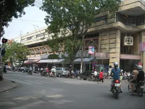 Bức ảnh chụp một góc phố đông đúc tại một thành phố, có khả năng là Hà Nội. Nổi bật trong ảnh là một tòa nhà lớn, nhiều tầng bằng bê tông, có kiến trúc giống một khu chợ hoặc trung tâm thương mại. Trên mặt tiền tòa nhà có một biểu tượng trang trí hình vuông với các vòng tròn đồng tâm bên trong. Tầng trệt của tòa nhà là một dãy các cửa hàng nhỏ có mái hiên.。Trên đường phố, dòng xe cộ tấp nập với rất nhiều xe máy và một vài chiếc ô tô. Hầu hết những người đi xe máy đều đội mũ bảo hiểm. Một số người đang dừng chờ đèn đỏ. Có cả người đi bộ trên vỉa hè.。Ven đường có cây xanh lớn, cột đèn giao thông và các biển báo. Có thể thấy một biển báo giao thông hình tròn viền đỏ, nền xanh với gạch chéo đỏ (biển báo cấm dừng và đỗ xe). Một số biển hiệu cửa hàng và biển quảng cáo cũng xuất hiện, trong đó có một tấm biển màu xanh ghi chữ "CÙNG CHIA SẺ" và một tấm áp phích màu hồng có hình một người phụ nữ. Một biển hiệu cửa hàng có chữ "San" cũng có thể được nhìn thấy.。（日本語訳）。写真は、ある都市の、おそらくハノイの、混雑した街角を撮影したものです。写真の中で目立っているのは、市場またはショッピングセンターのような建築様式の、大きなコンクリート製の多層階の建物です。建物のファサードには、内側に同心円を持つ四角形の装飾的なシンボルがあります。建物の1階は、ひさしのある小さな店舗が並んでいます。通りでは、非常に多くのバイクと数台の自動車で交通が混雑しています。バイクに乗っている人のほとんどはヘルメットを着用しています。何人かの人々は赤信号で停止して待っています。歩道には歩行者もいます。道端には大きな緑の木々、信号機の柱、そして標識があります。赤い縁取り、青い背景に赤い斜線が入った円形の交通標識（駐停車禁止の標識）が見えます。いくつかの店の看板や広告看板も見られ、その中には「CÙNG CHIA SẺ」と書かれた青い看板や、女性の絵が描かれたピンク色のポスターがあります。「San」という文字が入った店の看板も見ることができます。