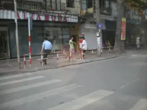 Đây là hình ảnh một con phố ở thành thị với các tòa nhà và cửa hàng. Ở phía bên kia đường, có một cửa hàng thời trang với biển hiệu lớn màu đỏ ghi chữ "ALTAMODA". Bên trong cửa kính có thể thấy một ma-nơ-canh đang mặc một chiếc váy màu trắng. Trên vỉa hè, một người đàn ông mặc áo sơ mi xanh nhạt đang ngồi trên lan can an toàn sơn sọc đỏ trắng. Gần đó, một nhóm thanh niên đang đứng nói chuyện. Ở phía xa bên phải, có một người đang đi bộ và đội nón lá. Dưới lòng đường có vạch kẻ sang đường dành cho người đi bộ. Treo trên một thân cây là một biểu ngữ dọc màu vàng có chữ đỏ nhưng không thể đọc rõ.。（日本語訳）。これは、ビルや店舗が立ち並ぶ都市の通りの画像です。道路の向こう側には、「ALTAMODA」と書かれた大きな赤い看板のあるファッション店があります。ガラス窓の内側には、白いドレスを着たマネキンが見えます。歩道では、水色のシャツを着た男性が、赤と白の縞模様に塗られた安全柵に座っています。その近くでは、若者の一団が立って話しています。右手の遠くには、歩いていてノンラーをかぶっている人がいます。道路には、歩行者用の横断歩道があります。木の幹には、赤い文字が書かれた黄色い縦長の横断幕が掛かっていますが、はっきりと読むことはできません。