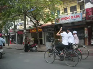 Bối cảnh là một con phố ở thành thị với các tòa nhà nhiều tầng san sát nhau và cây xanh được trồng ven đường. Trên đường, một nhóm ba người trẻ tuổi, có thể là học sinh mặc đồng phục áo trắng, đang đi xe đạp. Một người đang điều khiển xe và đưa tay lên vuốt tóc, một người khác ngồi ở yên sau. Bên lề đường, hai người phụ nữ đang đứng nói chuyện cạnh một chiếc xe máy màu đỏ. Khung cảnh cho thấy hoạt động sinh hoạt thường ngày trên một con phố ở Việt Nam, với người và xe cộ qua lại.。Các thông tin bằng chữ trên các biển hiệu của cửa hàng có thể đọc được bao gồm:。- Biển hiệu lớn màu xanh, chữ đỏ: "PHÒNG RĂNG". Bên dưới có các dòng chữ nhỏ hơn: "LONG NHA - 98 PHỐ HUẾ" và "LÂU NĂM - UY TÍN - CHẤT LƯỢNG".。- Biển hiệu bên cạnh: "HOÀNG ÁI VÂN ĐỒ HIỆU".。- Một cửa hàng khác có biển hiệu "ĐỒ HIỆU" và tên "... HIỆU" bị che một phần.。- Một cửa hàng có chữ "Cong audio".。（日本語訳）。背景は、多層階の建物が密集し、道沿いに街路樹が植えられている都市の通りです。路上では、白いシャツの制服を着た学生と思われる3人の若者のグループが自転車に乗っています。一人は自転車を運転し、手を上げて髪をかきあげており、もう一人は後ろのサドルに座っています。道端では、二人の女性が赤いバイクの隣で立って話しています。この光景は、人と乗り物が行き交う、ベトナムのある通りでの日常の生活活動を示しています。店の看板で読み取れる文字情報には以下が含まれます：。- 青地に赤文字の大きな看板：「歯科室」。その下にはより小さな文字の行があります：「LONG NHA - 98 PHỐ HUẾ」と「長年 - 信頼 - 品質」。- 隣の看板：「HOÀNG ÁI VÂN ブランド品」。- 別の店には「ブランド品」という看板があり、名前の「... HIỆU」は一部が隠れています。- 「Cong audio」という文字がある店。