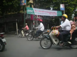 Bản ghi chữ viết trên băng rôn quảng cáo: 'ISC Học viện quản lý Singapore (SDC) Tel: 04. 2200184 Học viện PSB'. Ngoài ra còn có các biển hiệu khác nhưng chữ bị mờ, khó đọc.。Bối cảnh là một con đường nhựa trong thành phố với nhiều người và phương tiện qua lại. Hoạt động chính là mọi người đang điều khiển xe máy tham gia giao thông. Ở cận cảnh, một người đàn ông lớn tuổi mặc áo sơ mi trắng, đội mũ bảo hiểm màu vàng đang lái một chiếc xe máy. Phía trước ông có một bé gái mặc đồ màu hồng, đội mũ vải màu hồng. Một người khác ngồi sau lưng người đàn ông. Trên tay lái xe máy có treo một chiếc túi xách có hoa văn sặc sỡ. Phía sau họ là những người đi xe máy khác, bao gồm một người đàn ông đội mũ bảo hiểm màu đỏ và một người đàn ông khác mặc áo màu hồng. Dọc hai bên đường là các tòa nhà, cửa hàng và cây xanh.。（日本語訳）。広告バナーに書かれた文字の記録：「ISC シンガポール経営学院 (SDC) Tel: 04. 2200184 PSBアカデミー」。その他にも看板があるが、文字がかすれていて読みにくい。背景は、多くの人々や乗り物が行き交う都市のアスファルト道路である。主な活動は、人々がバイクを運転して交通に参加していることである。前景には、白いシャツを着て黄色いヘルメットをかぶった年配の男性がバイクを運転している。彼の前には、ピンク色の服を着てピンク色の布製の帽子をかぶった女の子がいる。もう一人が男性の後ろに乗っている。バイクのハンドルには、カラフルな模様のハンドバッグが掛けられている。彼らの後ろには他のバイクに乗る人々がおり、その中には赤いヘルメットをかぶった男性やピンク色のシャツを着た別の男性が含まれている。道路の両脇には、建物、店舗、そして緑の木々が並んでいる。