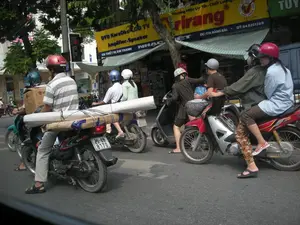 Bức ảnh chụp một cảnh giao thông đường phố ở Hà Nội. Nhiều người đang điều khiển xe máy và dừng lại trên đường, có thể là đang chờ đèn đỏ. Một người đàn ông lái xe máy đang chở hàng hóa cồng kềnh, bao gồm một thùng carton và một vật dài được cuộn lại buộc ở yên sau. Bên cạnh là một chiếc xe máy khác chở ba người. Hầu hết mọi người đều đội mũ bảo hiểm. Ở hậu cảnh là một cửa hàng bán thiết bị điện tử với tấm biển hiệu lớn màu vàng, trên đó có các dòng chữ: "Thương Hiệu Uy Tín - Chất Lượng", tên thương hiệu "Arirang", "DVD Karaoke LCD TV", "Amplifier-Speaker", "CH THIẾT BỊ ÂM THANH NHẠC". Địa chỉ và số điện thoại cũng được ghi rõ: "ĐC: 17A HÀNG BÀI" và "ĐT: 04 8251269". Ngoài ra còn có logo "Hàng Việt Nam chất lượng cao 2007".。（日本語訳）。写真はハノイの路上交通の一場面を撮影したものです。多くの人々がバイクを運転し、路上で停止しています。おそらく赤信号を待っているのでしょう。バイクを運転している一人の男性は、段ボール箱と、後部座席に縛り付けられた巻かれた長い物を含む、かさばる荷物を運んでいます。その隣には、3人乗りの別のバイクがいます。ほとんどの人がヘルメットを着用しています。背景には、大きな黄色い看板を掲げた電化製品店があり、そこには「Thương Hiệu Uy Tín - Chất Lượng」（信頼のブランド - 品質）、ブランド名の「Arirang」、「DVD Karaoke LCD TV」、「Amplifier-Speaker」、「CH THIẾT BỊ ÂM THANH NHẠC」（オーディオ・音楽機器店）といった文字が書かれています。住所と電話番号も「ĐC: 17A HÀNG BÀI」（住所：ハンバイ通り17A）と「ĐT: 04 8251269」（電話：04 8251269）と明記されています。その他に「Hàng Việt Nam chất lượng cao 2007」（2007年ベトナム高品質商品）のロゴもあります。