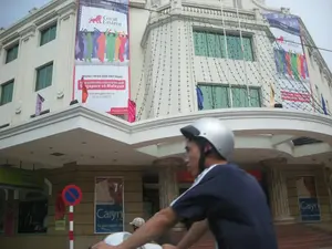 Bối cảnh là một con đường ở thành phố, phía trước một tòa nhà lớn, màu trắng theo kiến trúc hiện đại có các cột trụ. Một người đàn ông đội mũ bảo hiểm màu bạc đang điều khiển xe máy ở phía trước. Trên mặt tiền tòa nhà có treo nhiều biểu ngữ quảng cáo lớn.。Nội dung chữ viết trên các biểu ngữ và biển báo:。1. Biểu ngữ dọc lớn của công ty bảo hiểm 'Great Eastern': 'ARRIVED', 'Great Eastern', 'CHÚNG TÔI ĐÃ ĐẾN VIỆT NAM。', 'Công ty bảo hiểm nhân thọ lớn nhất Singapore và Malaysia', trang web 'www.lifeisgreat.com.vn', số điện thoại 'Tel: 84-4-9386757', và khẩu hiệu 'Cuộc Sống Tuyệt Vời'.。2. Áp phích ở tầng trệt: Quảng cáo sản phẩm 'Caryn' với dòng chữ 'Tã giấy người lớn'.。3. Biển báo giao thông: Một biển báo hình tròn viền đỏ, nền xanh có gạch chéo đỏ, là biển báo 'Cấm dừng và đỗ xe'.。Hoạt động chính được ghi lại là giao thông trên đường phố với người đi xe máy. Tòa nhà được trang trí để quảng bá cho sự kiện ra mắt của công ty Great Eastern tại Việt Nam. Ngoài ra còn có các dây đèn trang trí được treo trên mặt tiền của tòa nhà.。（日本語訳）。背景は都市の道路で、柱のある近代的な建築様式の大きな白い建物の前です。銀色のヘルメットをかぶった男性が、手前でバイクを運転しています。建物のファサードには、多くの大きな広告バナーが掲げられています。バナーと標識に書かれている文字の内容：。1. 保険会社「グレート・イースタン」の大きな縦長のバナー：「ARRIVED」、「Great Eastern」、「CHÚNG TÔI ĐÃ ĐẾN VIỆT NAM。」（私たちはベトナムに来ました。）、「シンガポールとマレーシアで最大の人生命保険会社」、ウェブサイト「www.lifeisgreat.com.vn」、電話番号「Tel: 84-4-9386757」、そしてスローガン「Cuộc Sống Tuyệt Vời」（素晴らしい人生）。2. 1階のポスター：「Caryn」製品の広告で、「Tã giấy người lớn」（大人用紙おむつ）という文字がある。3. 交通標識：赤い縁取りの円形で、青い背景に赤い斜線が入っており、「Cấm dừng và đỗ xe」（駐停車禁止）の標識です。記録されている主な活動は、バイクに乗る人がいる道路の交通です。建物は、グレート・イースタン社のベトナムでのローンチイベントを宣伝するために装飾されています。さらに、建物のファサードには装飾用の電飾も吊るされています。