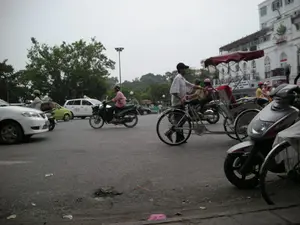 Đây là một cảnh đường phố tại một ngã tư hoặc bùng binh ở Hà Nội. Giao thông khá đông đúc với nhiều loại phương tiện. Ở phía trước, một người đàn ông đang đẩy một chiếc xích lô (cyclo), một loại xe ba bánh truyền thống. Xung quanh có nhiều xe máy đang lưu thông, người điều khiển và người ngồi sau đều đội mũ bảo hiểm. Ngoài ra còn có ô tô, bao gồm một chiếc taxi màu trắng có dòng chữ 'HANOI TAXI' và số điện thoại '3853 5353'. Ở phía bên phải là một tòa nhà lớn, màu trắng, có kiến trúc kiểu cổ. Trên tòa nhà có một số biển hiệu nhưng chữ viết khó đọc. Phía sau có cây cối xanh tươi và cột đèn đường. Bầu trời u ám, và mặt đường nhựa có một ít rác.。（日本語訳）。これはハノイの交差点またはロータリーでの街頭の風景です。交通は多くの種類の乗り物でかなり混雑しています。前方では、一人の男性が伝統的な三輪車であるシクロを押しています。周りには多くのバイクが通行しており、運転手も同乗者もヘルメットを着用しています。その他にも自動車があり、その中には「HANOI TAXI」という文字と電話番号「3853 5353」が書かれた白いタクシーも含まれています。右側には、古い様式の建築を持つ大きな白い建物があります。建物にはいくつかの看板がありますが、文字は読みにくいです。後方には青々とした木々と街灯があります。空は曇っており、アスファルトの路面には少しゴミがあります。