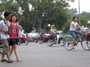 Bức ảnh chụp một cảnh sinh hoạt trên đường phố, có thể là ở Hà Nội, với nhiều người và phương tiện giao thông. Ở phía trước bên trái, một cặp đôi trẻ đang đi bộ. Người đàn ông mặc áo sơ mi trắng cộc tay, quần short màu be, cổ đeo một chiếc máy ảnh. Người phụ nữ đi cạnh mặc áo phông màu đỏ, chân váy ngắn màu đen và xách túi. Ở phía bên phải, một phụ nữ đang đi xe đạp. Trên đường có nhiều người khác đang đi xe máy, tất cả đều đội mũ bảo hiểm. Phía sau có một chiếc ô tô màu đen. Bối cảnh là một con đường nhựa ven một khu vực có nhiều cây cổ thụ xanh tốt, có thể là công viên hoặc bờ hồ. Không có văn bản hay biển hiệu nào có thể đọc được trong ảnh.。（日本語訳）。写真はおそらくハノイの、多くの人々や交通手段が行き交う路上での生活風景を捉えています。左手前には、若いカップルが歩いています。男性は白い半袖シャツにベージュのショートパンツを履き、首にはカメラをかけています。隣を歩く女性は赤いTシャツに黒いミニスカートを履き、バッグを持っています。右側には、一人の女性が自転車に乗っています。路上には他にもバイクに乗っている多くの人がおり、全員ヘルメットを着用しています。後ろには黒い車が一台あります。背景は、緑豊かな古い木々が多くある地域のそばを通るアスファルトの道で、公園か湖畔かもしれません。写真の中には、読み取れる文字や看板はありません。