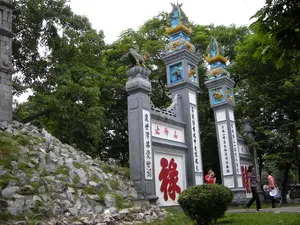 Bức ảnh chụp cổng vào của một khu di tích lịch sử hoặc đền chùa, có kiến trúc cổ kính. Đây là một chiếc cổng lớn với các cột trụ bằng xi măng, sơn màu xám. Trên đỉnh các cột trụ được trang trí công phu với các họa tiết rồng phượng màu xanh lam và vàng, cùng với tượng linh vật giống như con Nghê. Bên trái là một ngọn đồi nhỏ hoặc bờ kè được xây bằng đá và có cỏ mọc. Phía sau cổng là rất nhiều cây xanh tươi tốt. Một vài người dân đang đi bộ gần cổng, trong đó có một phụ nữ cầm nón lá, cho thấy hoạt động tham quan, vãn cảnh.。Nội dung chữ Hán trên cổng và các cột trụ được ghi lại như sau:。- Ba chữ phía trên cổng: "止仰山" (Chỉ Ngưỡng Sơn).。- Chữ lớn màu đỏ ở giữa: "祿" (Lộc).。- Câu đối trên các cột trụ (từ trái qua phải theo góc nhìn):。- Cột 1: "度世津梁覺世聞" (Độ thế tân lương giác thế văn).。- Cột 2: "尋派訪古此中來有燭光" (Tầm phái phỏng cổ thử trung lai hữu chúc quang).。- Cột 3: "慶成一塔五層聳翠聳層巒" (Khánh thành nhất tháp ngũ tầng tủng thúy tủng tầng loan).。- Cột 4: "表德垂功萬古一人傳聖德" (Biểu đức thùy công vạn cổ nhất nhân truyền thánh đức).。（日本語訳）。歴史的遺跡または寺院の入り口の門を撮影した写真で、古風な建築様式です。これはセメント製の柱を持つ大きな門で、灰色に塗られています。柱の上部は、青と金色の龍鳳の模様や、ゲ（Nghê）のような霊獣の像で精巧に装飾されています。左側には、石で造られ草が生えている小高い丘または土手があります。門の後ろには、青々と茂る多くの木々があります。門の近くを数人の人々が歩いており、その中にはノンラー（円錐形の葉笠）を持った女性もいて、観光や景色を楽しむ活動が見られます。門と柱に書かれた漢文の内容は以下の通りです。- 門の上部にある三文字：「止仰山」（Chỉ Ngưỡng Sơn）。- 中央にある大きな赤い文字：「祿」（Lộc）。- 柱の対聯（ついれん）（視点から見て左から右へ）：。- 柱1：「度世津梁覺世聞」（Độ thế tân lương giác thế văn）。- 柱2：「尋派訪古此中來有燭光」（Tầm phái phỏng cổ thử trung lai hữu chúc quang）。- 柱3：「慶成一塔五層聳翠聳層巒」（Khánh thành nhất tháp ngũ tầng tủng thúy tủng tầng loan）。- 柱4：「表德垂功萬古一人傳聖德」（Biểu đức thùy công vạn cổ nhất nhân truyền thánh đức）。