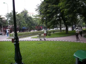 Bối cảnh là một công viên công cộng ở thành phố Hà Nội, với bãi cỏ xanh mướt, lối đi bộ lát gạch và nhiều cây xanh lớn. Ở phía trước là một cột đèn trang trí màu đen. Trên lối đi bộ, hai người đàn ông đang đi dạo. Phía bên kia công viên là một con phố tấp nập với nhiều phương tiện giao thông như ô tô và xe máy. Dọc theo con đường là các tòa nhà nhiều tầng với các cửa hàng ở tầng trệt. Có thể nhìn thấy một số biển hiệu, trong đó có một biển hiệu màu đỏ với chữ vàng ghi 'HÀ BÌNH'. Một bốt điện thoại công cộng cũng có thể được nhìn thấy bên lề đường. Khung cảnh mô tả một hoạt động thường ngày trong đô thị, với người dân đi dạo trong công viên và giao thông nhộn nhịp trên đường phố.。（日本語訳）。背景はハノイ市の公共の公園で、青々とした芝生、レンガ敷きの遊歩道、そして多くの大きな緑の木々があります。前方には黒い装飾的な街灯があります。遊歩道では、二人の男性が散歩しています。公園の向こう側は、自動車やバイクなどの多くの乗り物で賑わう通りです。道沿いには多層階の建物が並び、1階には店舗が入っています。いくつかの看板が見え、その中には「HÀ BÌNH」と金色の文字で書かれた赤い看板があります。道端には公衆電話ボックスも見えます。この光景は、公園を散歩する人々と路上のにぎやかな交通といった、都市における日常的な活動を描写しています。