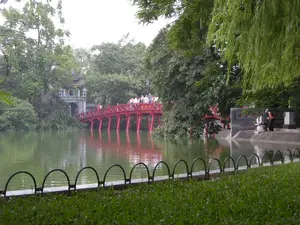 Bức ảnh ghi lại khung cảnh tại Hồ Hoàn Kiếm (Hồ Gươm) ở Hà Nội. Nổi bật trong ảnh là cây cầu Thê Húc sơn màu đỏ son, có kiến trúc cong bằng gỗ, bắc qua mặt hồ nước màu xanh lục để dẫn vào đền Ngọc Sơn. Trên cầu có nhiều người, có thể là du khách, đang đi bộ tham quan. Ở cuối cây cầu là cổng đền Ngọc Sơn, một công trình kiến trúc cổ với mái ngói. Trên cổng có các chữ Hán hoặc chữ Nôm nhưng không thể đọc rõ. Khung cảnh xung quanh được bao phủ bởi cây cối xanh tươi, bao gồm cả những cây liễu rủ bóng xuống mặt nước. Ở tiền cảnh là một bãi cỏ và hàng rào sắt thấp. Hoạt động chính diễn ra là tham quan, ngắm cảnh tại một địa danh lịch sử nổi tiếng.。（日本語訳）。写真はハノイのホアンキエム湖（ゴ・グオム湖）の風景を記録したものです。写真の中で目立つのは、朱色に塗られ、木造の湾曲した建築様式を持つテフック橋で、緑色の湖面に架かりゴックソン寺へと続いています。橋の上には多くの人がおり、観光客と思われ、歩いて見物しています。橋の終わりにはゴックソン寺の門があり、瓦屋根を持つ古い建築物です。門の上には漢字またはチュノムがありますが、はっきりと読むことはできません。周囲の風景は青々とした木々に覆われており、水面に影を落とす柳の木も含まれています。手前には芝生と低い鉄の柵があります。行われている主な活動は、有名な歴史的名所での観光、景色の鑑賞です。