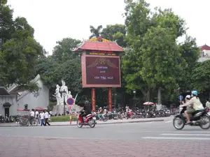 Bức ảnh chụp một khung cảnh đường phố tại Hà Nội. Ở trung tâm là một bảng điện tử lớn có mái che kiểu truyền thống, trên đó có các thông tin được ghi lại như sau: 'ĐẠI LỄ 1000 NĂM THĂNG LONG - HÀ NỘI', ngày '14/09/2008', giờ '14:51', và dòng chữ đếm ngược 'CÒN 756 NGÀY'. Bên trái bảng điện tử là một tượng đài bằng đá trắng, có thể là tượng đài 'Cảm tử cho Tổ quốc quyết sinh', với một nhóm các bức tượng người. Trên bệ tượng đài có khắc chữ nhưng khó đọc, chỉ nhận ra được vài từ như 'CẢM TỬ' và 'TỔ QUỐC'. Con đường phía trước có nhiều phương tiện qua lại, chủ yếu là xe máy. Nhiều người đang điều khiển xe máy trên đường, một số người đội mũ bảo hiểm. Ở bên lề đường, có nhiều xe máy khác đang đậu. Một nhóm thanh niên đang đi bộ trên vỉa hè gần tượng đài, và có một người đang đẩy một chiếc xe xích lô. Khung cảnh được bao quanh bởi nhiều cây xanh và các tòa nhà ở phía sau.。（日本語訳）。写真はハノイのある街角の風景を捉えたものです。中央には、伝統的な様式の屋根が付いた大きな電光掲示板があります。そこには次のような情報が記録されています。「タンロン・ハノイ遷都1000年大祝典」、日付「2008年9月14日」、時刻「14時51分」、そして「残り756日」というカウントダウンの文字です。電光掲示板の左側には白い石の記念碑があり、おそらくは「祖国のために決死」の記念碑で、一群の人々の像があります。記念碑の台座には文字が刻まれていますが、読み取りにくく、「CẢM TỬ（決死）」や「TỔ QUỐC（祖国）」といったいくつかの単語しか認識できません。手前の道路には多くの乗り物が行き交っており、主にバイクです。多くの人が道路でバイクを運転しており、何人かはヘルメットを着用しています。路肩には、他にも多くのバイクが駐車されています。記念碑の近くの歩道では若者の一団が歩いており、一人がシクロを押しています。風景は多くの緑の木々と、後方の建物に囲まれています。