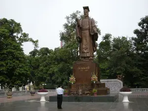 Trên bệ tượng có khắc dòng chữ: 'LÝ THÁI TỔ' và '974 - 1028'. Bức ảnh chụp một quảng trường công cộng ở Hà Nội, với trung tâm là tượng đài bằng đồng của Vua Lý Thái Tổ, người sáng lập kinh thành Thăng Long. Bức tượng được đặt trên một bệ đá lớn, uy nghi. Xung quanh là không gian rộng lớn được lát đá, bao bọc bởi nhiều cây xanh. Dưới chân tượng có đặt các lẵng hoa tươi, chậu cây cảnh và một lư hương, cho thấy đây là nơi tưởng niệm. Một người đàn ông mặc áo sơ mi trắng đang đứng trước tượng đài, có thể đang chụp ảnh hoặc bày tỏ lòng thành kính. Phía sau hàng cây có thể nhìn thấy một phần của một tòa nhà.。（日本語訳）。像の台座には「LÝ THÁI TỔ」と「974 - 1028」という文字が刻まれている。この写真はハノイのある公共広場を写したもので、その中心にはタンロン城を創建した李太祖（リー・タイ・トー）王の銅像がある。周囲は石畳の広大な空間で、多くの緑の木々に囲まれている。像の足元には生花のバスケット、鉢植えの観葉植物、そして香炉が置かれており、ここが記念の場所であることを示している。白いシャツを着た男性が像の前に立っており、写真を撮っているか、敬意を表しているのかもしれない。