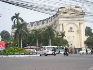 Bối cảnh là một góc phố đông đúc ở Hà Nội, nổi bật với một tòa nhà lớn có kiến trúc kiểu Pháp cổ. Tòa nhà này có màu vàng, trên tường có gắn logo và chữ 'Hilton' màu xanh. Trên đường phố, có nhiều phương tiện đang di chuyển, bao gồm ô tô, xe máy và một chiếc xe buýt màu trắng ở phía sau. Một chiếc taxi màu trắng có biển hiệu 'TAXI' trên nóc đang chạy trên đường. Nhiều người đang điều khiển xe máy, một số đội mũ bảo hiểm. Ở giữa đường có một dải phân cách được tạo bởi các khối bê tông sơn màu xanh và trắng với các mũi tên chỉ hướng. Xung quanh có trồng nhiều cây xanh, đặc biệt là cây cọ. Bầu trời phía trên chằng chịt dây điện và dây cáp. Ở phía bên trái có một tấm biển quảng cáo lớn màu đỏ, nhưng nội dung chữ viết không thể đọc rõ hoàn toàn.。（日本語訳）。背景はハノイの賑やかな街角で、古いフランス様式の建築を持つ大きな建物が際立っています。この建物は黄色で、壁には青い「Hilton」のロゴと文字が取り付けられています。路上では、多くの乗り物が移動しており、自動車、バイク、そして後方には一台の白いバスが含まれます。屋根に「TAXI」の表示がある白いタクシーが道を走っています。多くの人がバイクを運転しており、何人かはヘルメットを着用しています。道路の中央には、青と白に塗られ、方向を示す矢印が付いたコンクリートブロックでできた中央分離帯があります。周りには多くの緑の木々が植えられており、特にヤシの木があります。上空は電線やケーブルが張り巡らされています。
