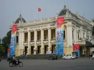 Bức ảnh chụp Nhà hát Lớn Hà Nội, một công trình kiến trúc lịch sử nổi tiếng.。**Chữ viết được ghi lại:**。*   Trên mặt tiền của tòa nhà có dòng chữ vàng khắc nổi: "NHÀ HÁT LỚN HÀ NỘI".。*   Hai bên lối vào chính có treo các băng rôn (phướn) dọc lớn để quảng bá cho một sự kiện. Các băng rôn có nội dung: "Vui Tết Trung Thu". Một băng rôn khác ghi "Tại Nhà Hát Lớn" và các thông tin liên quan đến chương trình, có thể nhận ra tên các đơn vị như Bộ Văn hóa, Du lịch và Thể thao và Đài Truyền hình Kỹ thuật số VTC.。**Mô tả cảnh quan và hoạt động:**。*   **Loại địa điểm:** Đây là Nhà hát Lớn Hà Nội, một nhà hát opera mang phong cách kiến trúc Pháp, nằm ở một quảng trường rộng ở trung tâm thành phố.。*   **Hoạt động:** Khung cảnh cho thấy hoạt động thường ngày tại khu vực này. Có người đang đi bộ trên các bậc thang của nhà hát và xung quanh quảng trường. Dưới lòng đường, có các phương tiện giao thông như ô tô và xe máy đang di chuyển. Một người đang lái xe máy chở người khác đi qua phía trước. Việc treo các băng rôn "Vui Tết Trung Thu" cho thấy không khí của dịp lễ hội Trung Thu.。*   **Vật thể:** Tòa nhà Nhà hát Lớn là đối tượng chính, với các cột trụ, ban công và mái vòm đặc trưng. Trên nóc tòa nhà có cột cờ treo lá cờ đỏ sao vàng của Việt Nam. Ngoài ra còn có các phương tiện giao thông như ô tô, xe máy và các băng rôn quảng cáo.。（日本語訳）。ハノイ・オペラハウスを撮影した写真。有名な歴史的建築物です。**記録されている文字：**。*   建物の正面には、「NHÀ HÁT LỚN HÀ NỘI」（ハノイ・オペラハウス）という金色の浮き彫り文字があります。*   正面玄関の両側には、イベントを宣伝するための大きな縦長の横断幕（幟）が掲げられています。横断幕には「Vui Tết Trung Thu」（楽しい中秋節）という内容が書かれています。別の横断幕には「Tại Nhà Hát Lớn」（オペラハウスにて）と書かれ、プログラムに関する情報が記載されており、文化・スポーツ・観光省やVTCデジタルテレビ局などの団体名が確認できます。**風景と活動の説明：**。*   **場所の種類：**これはハノイ・オペラハウスで、フランス建築様式のオペラハウスであり、市の中心部にある広い広場に位置しています。*   **活動：**この地域の日常的な活動がうかがえる風景です。劇場の階段や広場の周りを歩いている人々がいます。下の道路では、自動車やバイクなどの交通車両が移動しています。バイクを運転して他の人を乗せ、前を通り過ぎる人が一人います。*   **物体：**オペラハウスの建物が主要な対象物で、特徴的な柱、バルコニー、ドームがあります。建物の屋上には、ベトナムの金星紅旗を掲げた旗竿があります。その他にも、自動車、バイクなどの交通車両や広告の横断幕があります。