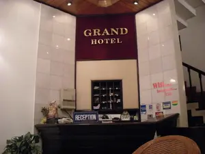 Đây là hình ảnh chụp quầy lễ tân của một khách sạn có tên là "GRAND HOTEL".。**Chữ viết được ghi lại:**。*   Trên tường phía sau quầy lễ tân có dòng chữ vàng "GRAND HOTEL" trên nền vải màu đỏ tía.。*   Trên quầy có một biển màu xanh lam với chữ trắng "RECEPTION".。*   Trên tường bên phải, có dòng chữ màu đỏ "Wifi... Internet... FREE".。*   Bên dưới dòng chữ Wifi là các nhãn dán chấp nhận thanh toán bằng thẻ tín dụng, bao gồm VISA, American Express, MasterCard, và các loại thẻ khác.。**Mô tả cảnh và vật thể:**。*   **Địa điểm:** Khu vực lễ tân của một khách sạn.。*   **Hoạt động:** Không có người và không có hoạt động nào đang diễn ra. Đây là một khung cảnh tĩnh.。*   **Vật thể:**。*   Quầy lễ tân được làm bằng gỗ tối màu.。*   Phía sau quầy có một tủ nhỏ có nhiều ngăn, có thể dùng để đựng chìa khóa hoặc thư từ.。*   Trên quầy có một hệ thống tổng đài điện thoại, một chiếc điện thoại bàn.。*   Một bức tượng nhỏ màu vàng, có thể là tượng Phật Di Lặc (Ông Địa), được đặt trên quầy cùng với một bình hoa nhỏ.。*   Tường được ốp bằng gạch men trắng bóng.。*   Ở phía sau bên phải, có thể nhìn thấy một phần của cầu thang bộ dẫn lên tầng trên.。*   Trần nhà có một phần được ốp gỗ và có đèn âm trần.。*   Ở phía trước bên trái có một chậu cây cảnh. Phía trước bên phải là một phần của chiếc ghế mây.。（日本語訳）。これは「GRAND HOTEL」という名前のホテルのフロントデスクを撮影した写真です。**記録されている文字：**。*   フロントデスクの後ろの壁には、深紅色の布地を背景に「GRAND HOTEL」という金色の文字があります。*   カウンターの上には、「RECEPTION」と白文字で書かれた青い看板があります。*   右側の壁には、「Wifi... Internet... FREE」という赤い文字があります。*   Wifiの文字の下には、VISA、American Express、MasterCard、その他のカードを含む、クレジットカードでの支払いを受け付けるステッカーがあります。**風景と物体の説明：**。*   **場所：** ホテルのフロントエリア。*   **活動：** 人はおらず、何の活動も行われていません。これは静的な光景です。*   **物体：**。*   フロントデスクはダークカラーの木材で作られています。*   カウンターの後ろには、鍵や手紙を保管するために使用できる、多くの仕切りがある小さな棚があります。*   カウンターの上には、電話交換システム、卓上電話機があります。*   小さな金色の像、おそらく弥勒菩薩（布袋様）の像が、小さな花瓶と一緒にカウンターの上に置かれています。*   壁は光沢のある白いセラミックタイルで覆われています。*   右奥には、上の階へ続く階段の一部が見えます。*   天井の一部は木で覆われており、埋め込み式の照明があります。*   左手前には観葉植物の鉢があります。右手前には籐の椅子の一部があります。