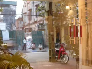 Bức ảnh chụp một khung cảnh đường phố ở Hà Nội, nhìn từ bên trong một tòa nhà qua một cánh cửa kính. Ở phía trước, một chiếc xe máy màu đỏ đang đỗ trên vỉa hè, bên cạnh một cột điện. Cánh cửa kính có tay nắm hiện đại màu đỏ và vàng. Bên kia đường, có một hàng rào bằng tôn lượn sóng màu xanh lá cây, có thể là một công trường xây dựng. Trên hàng rào có dán các tờ giấy thông báo màu trắng nhưng không thể đọc rõ nội dung. Có ba người đàn ông đang ở trên vỉa hè; một người đang đứng và hai người đang ngồi xổm, có vẻ như họ đang nghỉ ngơi. Phía sau hàng rào là những ngôi nhà phố cũ, nhiều tầng, mang nét kiến trúc cổ điển.。（日本語訳）。ハノイのある街の風景を、建物の中からガラスのドアを通して撮影した写真です。手前には、歩道に赤いバイクが電柱の隣に停まっています。ガラスのドアには、赤と黄色のモダンな取っ手が付いています。道の向こう側には、緑色の波板でできた柵があり、おそらく工事現場でしょう。柵には白いお知らせの紙が貼られていますが、内容ははっきりと読み取れません。歩道には3人の男性がいます。1人は立っており、2人はしゃがんでいて、休憩しているようです。柵の後ろには、古典的な建築様式を持つ、多層階の古いタウンハウスが建っています。
