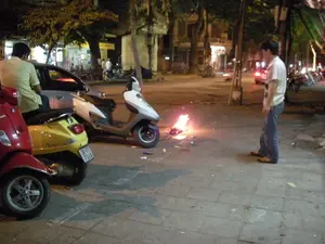 Bối cảnh là một con phố ở Hà Nội vào ban đêm. Trên vỉa hè lát gạch, một người đàn ông mặc áo phông trắng và quần jean đang đứng nhìn một đống lửa nhỏ đang cháy. Đây dường như là hoạt động đốt vàng mã, một nghi lễ tín ngưỡng. Gần đó, có nhiều xe máy đang đỗ, bao gồm một chiếc xe tay ga màu đỏ, một chiếc Vespa màu vàng và một chiếc xe màu trắng. Một người đàn ông khác mặc áo phông kẻ đang ngồi trên chiếc xe Vespa màu vàng. Một chiếc ô tô màu bạc cũng đang đỗ bên cạnh. Phía sau là khung cảnh đường phố với cây cối, các tòa nhà và ánh đèn từ các cửa hàng. Có thể nhìn thấy một vài biển hiệu nhưng chữ trên đó không thể đọc rõ.。（日本語訳）。背景は夜のハノイのある通りです。レンガ敷きの歩道の上で、白いTシャツとジーンズを履いた男性が、燃えている小さな火の山を立って見ています。これは、宗教的な儀式である冥銭を燃やす行為のようです。その近くには、赤いスクーター、黄色いベスパ、そして白いバイクを含む多くのバイクが駐車しています。縞模様のTシャツを着た別の男性が、黄色いベスパに座っています。銀色の自動車も隣に駐車しています。後方には、木々、建物、そして店からの明かりがある街の風景が広がっています。いくつかの看板が見えますが、その上の文字ははっきりと読み取れません。