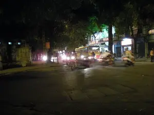 Đây là một cảnh đường phố ở Hà Nội vào ban đêm. Con đường có nhiều phương tiện giao thông đang di chuyển, chủ yếu là xe máy với đèn pha đang bật sáng. Do ảnh bị mờ vì chuyển động nên hình ảnh các phương tiện và người lái không rõ nét. Ở phía sau có một chiếc xe buýt màu vàng. Hai bên đường là những hàng cây lớn. Phía bên phải là các tòa nhà có cửa hàng đang sáng đèn. Có một tấm biển hiệu lớn được chiếu sáng với các chữ màu đỏ nhưng không thể đọc rõ nội dung. Cảnh tượng cho thấy hoạt động giao thông về đêm trên một con phố của thành phố.。（日本語訳）。これは夜のハノイの街頭の風景です。道路には多くの交通車両が移動しており、主にヘッドライトを点灯させたバイクです。写真は動きによってぼやけているため、車両や運転手の姿ははっきりしません。後方には黄色いバスが1台あります。道路の両側には大きな並木があります。右側には、明かりが灯っている店舗のある建物があります。赤い文字で照らされた大きな看板がありますが、内容ははっきりと読み取れません。この光景は、ある都市の通りでの夜間の交通状況を示しています。