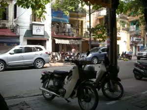 Bối cảnh là một con phố ở Hà Nội với những tòa nhà ống cao tầng đặc trưng san sát nhau. Dọc theo con đường và trên vỉa hè, có nhiều phương tiện giao thông đang đỗ, bao gồm cả ô tô và xe máy. Nổi bật ở phía trước là hai chiếc xe máy, và bên đường là một chiếc ô tô màu bạc. Trên các tòa nhà có một số biển hiệu. Một biển hiệu màu trắng có mái che nhỏ màu xanh lá cây ghi dòng chữ "CÔNG TY TNHH THƯƠNG MẠI" và các ký tự chữ Hán "和宝堂". Một mái hiên gần đó có logo của hãng bia "Tiger". Ở phía xa, có một người đang đi bộ và một người khác dường như đang bán hàng trên vỉa hè. Cây cối hai bên đường tỏa bóng mát, tạo nên một khung cảnh đường phố điển hình của một ngày ở Hà Nội.。（日本語訳）。背景はハノイのある通りで、特徴的な高層のチューブハウスが隣接して建ち並んでいます。道路沿いや歩道には、自動車やバイクを含む多くの交通手段が駐車しています。前景には2台のバイクが目立っており、道路脇には銀色の自動車が1台あります。建物にはいくつかの看板があります。小さな緑色の庇が付いた白い看板には、「CÔNG TY TNHH THƯƠNG MẠI」という文字と「和宝堂」という漢字が書かれています。その近くの軒先には、ビールブランド「Tiger」のロゴがあります。道路の両側の木々が涼しい影を落とし、ハノイのある日の典型的な街の風景を作り出しています。