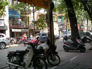 Đây là một khung cảnh đường phố tại Hà Nội. Con phố có nhiều cây xanh lớn, vỉa hè lát gạch và các tòa nhà ống nhiều tầng san sát nhau. Rất nhiều xe máy, bao gồm cả xe số và xe tay ga, đang được dựng trên vỉa hè và dưới lòng đường, bên cạnh một vài chiếc ô tô. Một người đàn ông đội mũ bảo hiểm đang điều khiển xe máy trên đường, trong khi một nhóm phụ nữ đang đi bộ trên vỉa hè. Có thể thấy nhiều biển hiệu cửa hàng: một biển hiệu ghi "CẮT TÓC NAM NỮ", một biển hiệu khác ở xa hơn có chữ "TÓC THỜI TRANG" và "INTERNET". Trên một tòa nhà có một tấm biển quảng cáo lớn màu xanh lam nhưng chữ viết không rõ ràng. Trên một cột điện có dán một tờ giấy quảng cáo màu vàng với số "568". Khung cảnh thể hiện nhịp sống sinh hoạt thường ngày trên một con phố đặc trưng của Việt Nam.。（日本語訳）。これはハノイのある通りの風景です。通りには大きな緑の木々が多く、歩道はタイルで舗装され、多層階のチューブハウスが密集しています。カブ型バイクやスクーターを含む多くのバイクが、数台の自動車の隣で、歩道や車道に停められています。ヘルメットをかぶった男性が路上でバイクを運転しており、一方、女性のグループが歩道を歩いています。多くの店の看板が見えます。ある看板には「CẮT TÓC NAM NỮ」（男女のヘアカット）、遠くにある別の看板には「TÓC THỜI TRANG」（ファッションヘア）と「INTERNET」の文字があります。ある建物には大きな青い広告看板がありますが、文字ははっきりしません。電柱には「568」という数字が書かれた黄色い広告紙が貼られています。この風景は、ベトナムの典型的な通りにおける日常の生活のリズムを表しています。