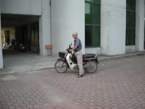 Bức ảnh chụp một người đàn ông lớn tuổi, tóc bạc, mặc áo sơ mi cộc tay màu xanh và quần dài màu be, đang đứng cạnh một chiếc xe máy số. Chiếc xe máy có màu tím và trắng, phía trước có gắn một chiếc giỏ đen. Người đàn ông đang nhìn về phía người chụp ảnh. Bối cảnh là một khoảng sân được lát gạch block màu đỏ, phía trước một tòa nhà lớn, hiện đại. Tòa nhà có tường sơn trắng và các cửa sổ lớn bằng kính màu xanh lục. Bên trái là một lối vào có mái che, giống như một nhà để xe, nơi có một chiếc xe máy khác và một chiếc xe đạp đang đỗ. Gần lối vào có một biển báo nhỏ màu cam, chữ đỏ. Chữ trên biển báo bị mờ và không thể đọc rõ, nhưng có khả năng đây là biển chỉ dẫn khu vực đỗ xe.。（日本語訳）。彼は青い半袖シャツとベージュの長ズボンを着用し、スクーターの隣に立っています。スクーターは紫色と白色で、前には黒いカゴが取り付けられています。男性は撮影者の方を見ています。背景は、大きくて近代的な建物の前にある、赤いブロックタイルで舗装された庭です。建物は白い壁と緑色のガラスの大きな窓があります。左側には屋根付きの入り口があり、ガレージのようです。そこには別のバイクと自転車が停まっています。入り口の近くには、オレンジ色に赤い文字の小さな看板があります。看板の文字はぼやけていてはっきりと読むことはできませんが、おそらく駐車エリアを示す案内板でしょう。