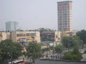 Bức ảnh chụp một góc cảnh quan đô thị ở Hà Nội, có thể vào một ngày trời nhiều sương mù. Khung cảnh cho thấy sự pha trộn giữa các công trình kiến trúc cũ và mới. Nổi bật là một tòa nhà chung cư cao tầng hiện đại, sơn màu be và hồng. Xung quanh nó là các khu nhà tập thể cũ, thấp tầng hơn, với các đặc điểm quen thuộc như 'chuồng cọp' (lồng sắt cơi nới ban công), nhiều cục nóng điều hòa và các bồn chứa nước bằng inox trên mái. Phía xa, hai tòa nhà cao tầng khác có màu xanh lục mờ ảo trong sương. Dưới mặt đất là một con đường đông đúc với nhiều phương tiện giao thông, bao gồm rất nhiều xe máy, ô tô và một chiếc xe buýt. Có thể thấy người dân đang tham gia giao thông, một số đang dừng chờ ở cột đèn tín hiệu. Cây xanh được trồng nhiều dọc hai bên đường và xen kẽ giữa các tòa nhà. Có một số biển quảng cáo ở tầng trệt, trong đó có một biển hiệu màu vàng có thể đọc được chữ 'FINATEX' và một biển hiệu khác có chữ 'PROSTEE...'. Các biển quảng cáo lớn khác thì không thể đọc rõ chữ.。（日本語訳）。この写真はハノイの都市景観の一角を撮影したもので、おそらく霧の深い日だと思われます。この風景は、新旧の建築物が混在している様子を示しています。目立っているのは、ベージュとピンク色に塗られた近代的な高層マンションです。その周りには、より低層の古い集合住宅があり、「虎の檻」（バルコニーを拡張した鉄製のケージ）、多くのエアコン室外機、屋上のステンレス製貯水タンクといったおなじみの特徴が見られます。遠くには、他の2つの高層ビルが霧の中に緑色がかってぼんやりと見えます。地上には交通量の多い道路があり、非常に多くのバイク、自動車、そして1台のバスを含む多くの交通手段が見られます。人々が通行しているのが見え、一部は信号機で停止して待っています。道路の両脇や建物の間に多くの緑の木々が植えられています。1階にはいくつかの広告看板があり、その中には「FINATEX」という文字が読み取れる黄色い看板と、「PROSTEE...」という文字がある別の看板があります。他の大きな広告看板は文字がはっきりと読み取れません。