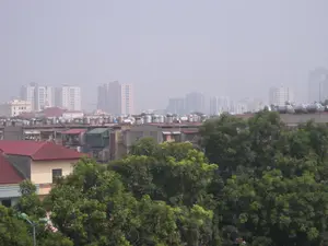 Bức ảnh chụp một quang cảnh đô thị, có thể là Hà Nội, nhìn từ trên cao. Bầu trời bị bao phủ bởi một lớp sương mù dày đặc, làm giảm tầm nhìn. Ở phía xa là nhiều tòa nhà chung cư cao tầng hiện đại mờ ảo trong sương. Ở khu vực giữa và gần hơn là các tòa nhà thấp tầng, kiểu nhà tập thể hoặc nhà riêng cũ hơn. Một đặc điểm nổi bật là trên sân thượng của các tòa nhà này có rất nhiều bồn chứa nước bằng inox và các hệ thống máy nước nóng năng lượng mặt trời. Phía trước là những tán cây xanh tươi tốt, che khuất một phần các ngôi nhà ở tầng thấp hơn. Không có chữ viết nào có thể đọc được. Khung cảnh cho thấy sự pha trộn giữa kiến trúc cũ và mới trong một thành phố đang phát triển, với dấu hiệu của ô nhiễm không khí hoặc sương mù.。（日本語訳）。写真は、おそらくハノイの都市景観を上から撮影したものです。空は厚い霧に覆われ、視界が悪くなっています。遠方には、多くの近代的な高層マンションが霧の中にぼんやりと見えます。中景から近景にかけては、低層の建物、集合住宅タイプ、またはより古い個人住宅があります。際立った特徴は、これらの建物の屋上に非常に多くのステンレス製貯水タンクと太陽熱温水器システムがあることです。手前には青々とした木々の梢があり、下層階の家々を部分的に覆い隠しています。読み取れる文字はありません。この光景は、発展途上の都市における新旧の建築様式の混在と、大気汚染または霧の兆候を示しています。