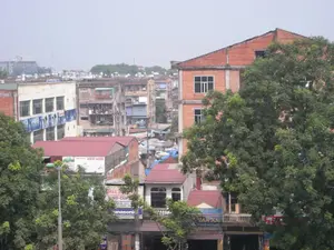 Đây là quang cảnh một khu dân cư đô thị đông đúc ở Hà Nội, được chụp từ trên cao nhìn xuống. Khung cảnh bao gồm nhiều tòa nhà cao tầng san sát nhau với kiến trúc đa dạng, từ những khu tập thể cũ đến những ngôi nhà mới xây bằng gạch đỏ. Cây cối xanh tươi mọc um tùm xen kẽ giữa các công trình. Ở phía xa, trên nóc các tòa nhà có thể thấy nhiều bồn chứa nước. Phía dưới, giữa các tòa nhà, có một khu vực trông giống như một khu chợ với các mái che bằng bạt màu xanh. Không có hoạt động cụ thể nào của con người được thể hiện rõ, nhưng sự hiện diện của các biển hiệu cho thấy đây là một khu vực có hoạt động thương mại. Các chữ viết trên biển hiệu có thể nhận dạng được bao gồm: 'DUY ANH', 'SINO-CLIP', 'Panasonic', và 'VAN AN'. Ngoài ra còn có các biển hiệu và băng rôn khác nhưng chữ bị mờ, không thể đọc rõ.。（日本語訳）。これはハノイのある密集した都市部の住宅地の光景で、上から見下ろすように撮影されたものです。この光景には、古い集合住宅から赤レンガで新築された家まで、多様な建築様式の高層ビルが数多く密集しています。建物の間には、青々とした木々が生い茂っています。遠くの建物の屋上には、多くの貯水タンクが見えます。下の方、建物の間には、青い防水シートの屋根がある市場のようなエリアがあります。特定の人間の活動ははっきりと見えませんが、看板の存在は、ここが商業活動のある地域であることを示しています。認識可能な看板の文字には、「DUY ANH」、「SINO-CLIP」、「Panasonic」、「VAN AN」が含まれます。その他にも看板や横断幕がありますが、文字がぼやけていてはっきりと読むことはできません。