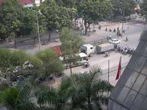 Bức ảnh được chụp từ trên cao, cho thấy một khung cảnh giao thông tại một ngã tư đường phố đông đúc ở một thành phố, có thể là Hà Nội. Con đường được trải nhựa và có nhiều phương tiện qua lại.。Hoạt động chính là giao thông. Rất nhiều người đang điều khiển xe máy, nhiều người trong số họ đội mũ bảo hiểm. Một nhóm người đi xe máy đang dừng lại ở ngã tư, có lẽ đang chờ đèn tín hiệu. Một chiếc xe tải lớn màu trắng đang kéo rơ moóc chở hàng và một chiếc ô tô con màu trắng cũng đang di chuyển trong khu vực ngã tư. Một người đi bộ đang băng qua đường. Dưới bóng cây bên lề đường, có một số xe máy đang đỗ.。Các vật thể có trong ảnh bao gồm nhiều loại phương tiện như xe máy, ô tô con, xe tải, xe đầu kéo và một chiếc xe khách nhỏ màu trắng. Có một thùng container màu đỏ, trên đó có chữ trắng nhưng không thể đọc rõ. Hai bên đường được trồng rất nhiều cây xanh lớn, tán lá rậm rạp. Phía trước có một vài cây cọ. Có thể thấy các cột đèn đường, dây điện và một lá cờ đỏ sao vàng (quốc kỳ Việt Nam) đang được treo trên cột. Ở phía trước bên phải là một phần của mái nhà bằng kính.。（日本語訳）。写真は上から撮影されたもので、ある都市、おそらくハノイの、混雑した街の交差点の交通風景を写している。道路はアスファルトで舗装されており、多くの車両が往来している。主な活動は交通である。非常に多くの人々がバイクを運転しており、その多くはヘルメットを着用している。バイクに乗った一団が交差点で停止しており、おそらく信号を待っている。一台の大きな白いトラックが貨物を積んだトレーラーを牽引しており、一台の白い乗用車も交差点内を移動している。一人の歩行者が道路を横断している。道端の木陰に、数台のバイクが駐車している。写真に写っている物体には、バイク、乗用車、トラック、トレーラーヘッド、そして一台の白い小型バスといった多くの種類の車両が含まれる。赤いコンテナが一つあり、その上には白い文字があるが、はっきりと読むことはできない。道路の両側には、葉が鬱蒼と茂った大きな緑の木がたくさん植えられている。前方には数本のヤシの木がある。右手前にはガラスの屋根の一部がある。