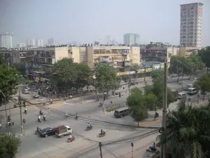 Đây là một bức ảnh chụp từ trên cao, bao quát một ngã tư đường phố sầm uất tại một khu đô thị, có khả năng là Hà Nội. Bối cảnh cho thấy sự pha trộn giữa kiến trúc cũ và mới. Ở hậu cảnh và hai bên là những khu nhà tập thể cũ kỹ, nhiều tầng, đặc trưng bởi các ban công được cơi nới ("chuồng cọp"). Xen kẽ là các tòa nhà cao tầng hiện đại hơn mọc lên ở phía xa, nổi bật là một tòa nhà cao tầng ở phía bên phải. Hoạt động chính trong ảnh là giao thông đông đúc. Rất nhiều người đang điều khiển xe máy, phương tiện phổ biến nhất. Ngoài ra còn có ô tô, một chiếc xe tải nhỏ màu trắng, một chiếc xe buýt nhỏ màu trắng sọc xanh và một vài người đi xe đạp. Mọi người đang di chuyển qua lại ngã tư, tạo nên một khung cảnh nhộn nhịp đặc trưng của đô thị Việt Nam. Dọc các con đường và trên dải phân cách có nhiều cây xanh. Hệ thống dây điện và cáp viễn thông chằng chịt vắt ngang phía trên. Trên các tòa nhà có một số biển quảng cáo, tuy nhiên chữ viết trên đó quá mờ để có thể đọc rõ, chỉ có thể nhận ra một biển hiệu màu vàng có chữ "VINA...".。（日本語訳）。これは上空から撮影された写真で、都市部、おそらくハノイの賑やかな交差点を捉えています。背景には、新旧の建築が混在している様子が見られます。背景と両側には、増築されたバルコニー（「虎の檻」）が特徴的な、古くて多層の集合住宅があります。その間に、遠くにそびえ立つより近代的な高層ビルが点在しており、特に右側にある高層ビルが目立ちます。写真の中の主な活動は、混雑した交通です。非常に多くの人々が、最も一般的な乗り物であるバイクを運転しています。その他にも、自動車、白い小型トラック1台、白地に青い縞模様の小型バス1台、そして数人の自転車に乗っている人がいます。人々は交差点を行き交い、ベトナムの都市に特有の賑やかな光景を作り出しています。道路沿いや中央分離帯には多くの緑の木々があります。頭上には、電線や通信ケーブルが複雑に張り巡らされています。建物にはいくつかの広告看板がありますが、その上の文字はぼやけすぎてはっきりと読むことはできず、「VINA...」という文字のある黄色い看板が一つ認識できるだけです。